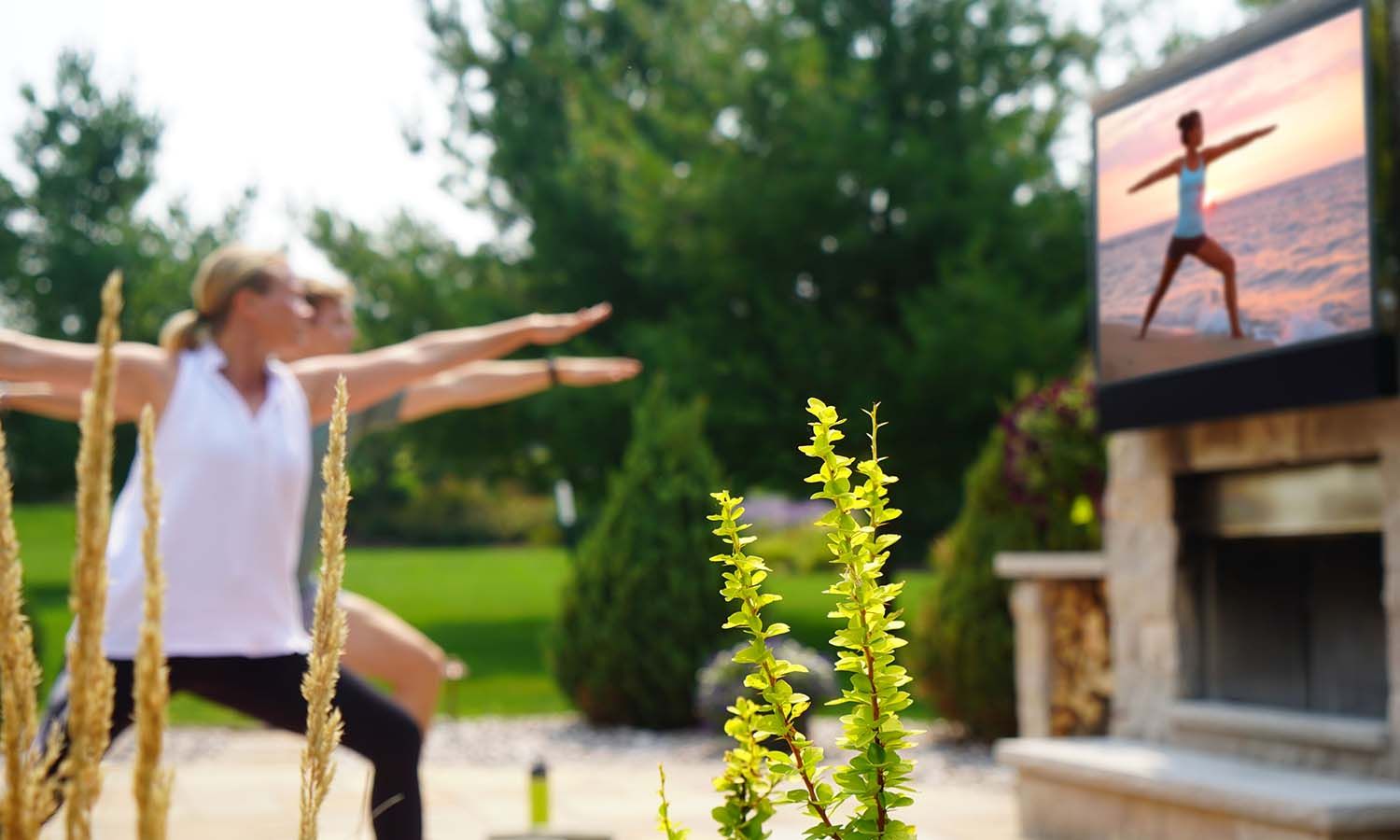 A group of people practicing yoga outdoors on a patio, following along with a fitness video playing on a mounted TV above an outdoor fireplace, surrounded by greenery.